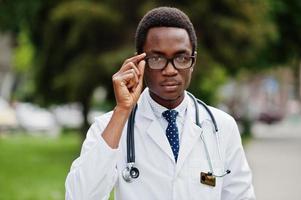 elegante médico afroamericano con estetoscopio y bata de laboratorio, con anteojos posados al aire libre. foto