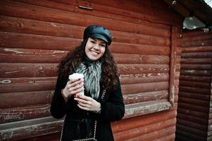 chica mexicana rizada con gorra de cuero y taza de café de plástico a mano contra el mercado de madera. foto