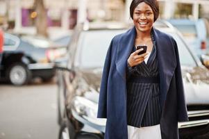éxito elegante mujer afroamericana en abrigo contra coche suv de negocios negro mirando el teléfono.