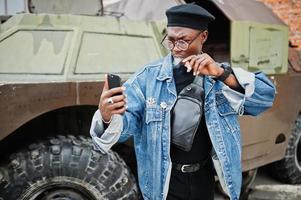 African american man in jeans jacket, beret and eyeglasses, smoking cigar and posed against btr military armored vehicle, making selfie on phone. photo
