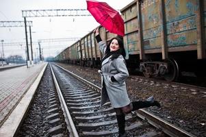 chica morena con abrigo gris con paraguas rojo en la estación de tren. foto