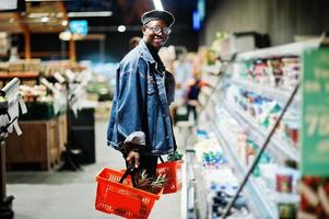 elegante hombre afroamericano casual con chaqueta de jeans y boina negra sosteniendo dos canastas, parado cerca de la nevera y comprando en el supermercado. foto