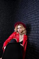 Studio portrait of blonde girl in red hat, glasses and leather jacket posed on chair against brick wall. photo