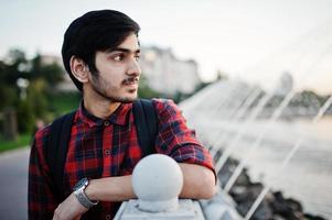 Young indian student man at checkered shirt and jeans with backpack posed on evening city against fountains. photo