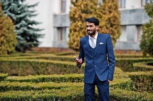 hombre indio de barba elegante con bindi en la frente, vestido con traje azul posado al aire libre contra arbustos verdes en el parque. foto