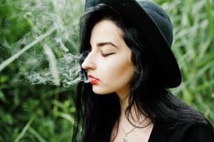 chica fumadora sensual todo en negro, labios rojos y sombrero. mujer gótica dramática fumando en caña común. foto
