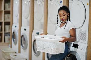 alegre mujer afroamericana con cesta blanca cerca de la lavadora en la lavandería de autoservicio.