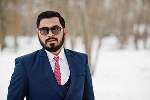 primer plano retrato de elegante hombre de negocios de barba india en traje y gafas de sol posado en el día de invierno al aire libre. foto