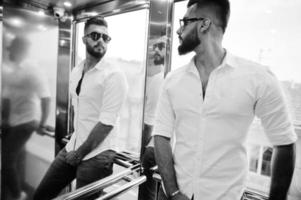 Stylish tall arabian man model in white shirt and sunglasses posed at elevator inside. photo