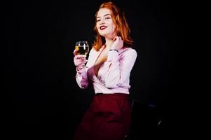 Gorgeous red haired girl in pink blouse and red skirt with glass of wine at hand isolated on black. photo