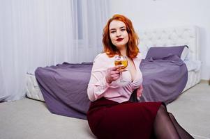 Gorgeous red haired girl in pink blouse and red skirt with glass of wine at hand near bed at room. photo