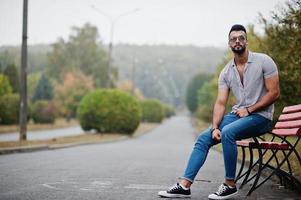 Fashionable tall arab beard man wear on shirt, jeans and sunglasses posed on park at bench. photo