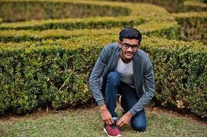 Stylish indian man at glasses wear casual posed outdoor and tie the laces on the sneakers. photo