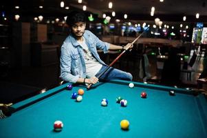 Stylish asian man wear on jeans playing pool billiard on bar. photo