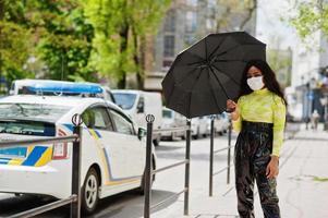 African American woman posing with facial mask to protect from infections from bacteria, viruses and epidemics and hold umbrella against police car. photo