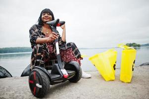 hermosa mujer afroamericana sentada cerca de segway o hoverboard. chica negra con símbolo de reciclaje de bolsas ecológicas de tela amarilla. foto