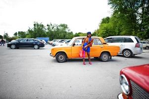 hermosa dama afroamericana con bolsas de compras de pie cerca de un auto retro clásico naranja. foto