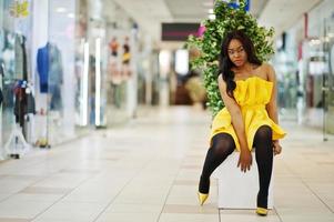 Stylish african american woman at yellow dreess posed at mall and sitting on bench inside. photo