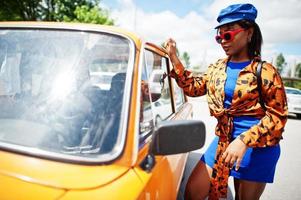 hermosa dama afroamericana con gafas de sol de pie cerca de un auto retro clásico naranja. foto