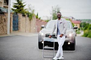 Rich and stylish african american man in blazer and white pants, eyeglasses posed against suv car. photo