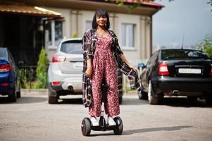 hermosa mujer afroamericana usando segway o hoverboard. chica negra en scooter eléctrico autoequilibrado de doble rueda contra estacionamiento. foto