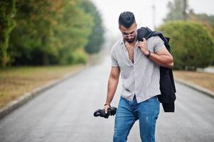 Fashionable tall arab beard man wear on shirt, jeans and sunglasses walking at park with umbrella and coat at hand. photo