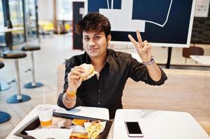 Stylish indian man sitting at fast food cafe and eating hamburger and gesture peace sign hand. photo