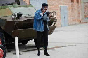 African american man in jeans jacket, beret and eyeglasses, with cigar posed against btr military armored vehicle, and speaking on mobile phone. photo