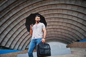 el hombre de barba árabe alto de moda usa camisa, jeans y gafas de sol con paraguas y bolso de caja posado en el salón de la arena. foto