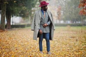 elegante modelo afroamericano con abrigo gris, corbata de chaqueta y sombrero rojo posado en la calle con niebla en otoño. foto