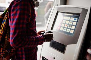 hombre afroamericano en camisa a cuadros con mochila sosteniendo billetera en mano contra atm. viajero hombre negro. foto