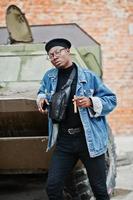 African american man in jeans jacket, beret and eyeglasses, with cigar posed against btr military armored vehicle. photo