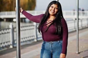 Pretty latino xxl model girl from Ecuador wear on violet blouse posed against lake. photo