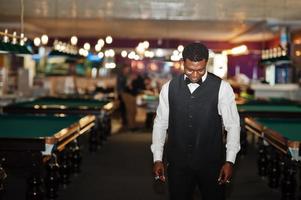 joven y apuesto hombre africano con camisa blanca, chaleco negro y corbata de moño frente a mesas de billar. foto