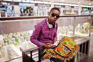 hombre afroamericano con camisa a cuadros, gafas de sol y jeans con maleta y mochila. el viajero negro revisó su pasaporte en la tienda libre de impuestos. foto