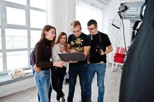 Photographer explaining about the shot to his team in the studio and looking on laptop. Talking to his assistants holding a camera during a photo shoot. Teamwork and brainstorm.