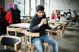Casual and stylish young asian man with earphones at cafe drinking juice from glass and holding tablet. photo