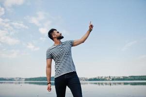 Handsome tall arabian beard man model at stripped shirt posed outdoor against lake and showing finger into the sky. Fashionable arab guy. photo