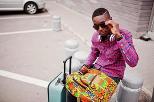 African american man in checkered shirt, sunglasses and earphones with suitcase and backpack. Black man traveler holding mobile phone at hand. photo