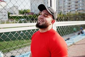 joven musculoso barbudo brutal vestido con camisa roja, pantalones cortos y gorra en el estadio. foto