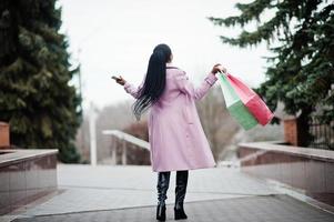 joven y elegante mujer afroamericana en la calle, vestida con un abrigo de moda con bolsas de compras y teléfono móvil en las manos. foto