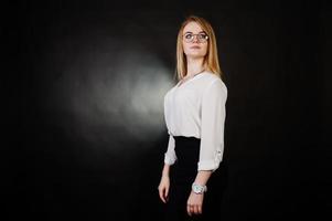 Studio portrait of blonde businesswoman in glasses, white blouse and black skirt against dark background. Successful woman and stylish girl concept. photo