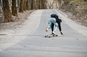 fallar al caer de una patineta. hombre árabe de estilo callejero en anteojos con longboard longboarding por el camino. foto