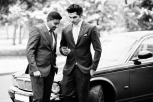 Two elegant indian fashionable mans model on suit posed at winter day against black classic car and looking at phone. photo