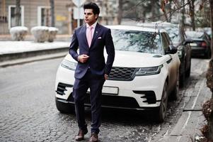 elegante modelo de hombre macho indio con traje y corbata rosa posado contra un coche de negocios blanco. foto