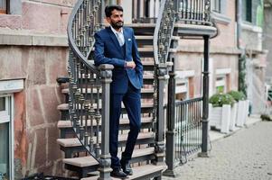 hombre indio de barba elegante con bindi en la frente, vestido con traje azul posado al aire libre contra escaleras de hierro. foto