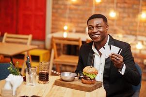 Respectable young african american man in black suit sitting in restaurant hold a lot of credit cards with tasty double burger and soda drink. photo