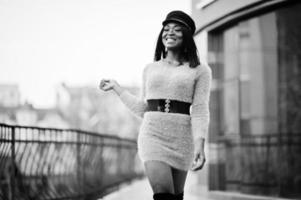 African american woman at violet dress and cap posed outdoor against modern building. photo