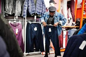 Stylish casual african american man at jeans jacket and black beret at clothes store looking on new sport pants. photo