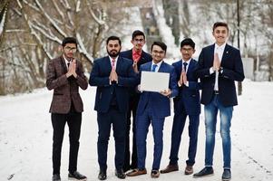 grupo de seis hombres de negocios indios en trajes posaron al aire libre en un día de invierno en europa, mirando en la computadora portátil y mostrando las manos de namaste. foto